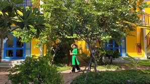 A person walking in a courtyard with a yellow building, surrounded by greenery and a wooden deck. - Chez Mimosa Hoi An - Rice Field Retreat (Da Nang)