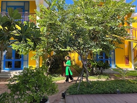 Una persona caminando en un patio con un edificio amarillo, rodeado de follaje y una terraza de madera.