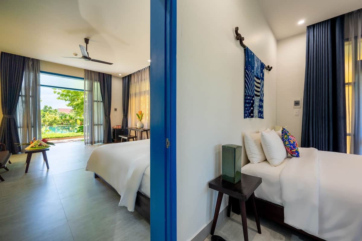 A hotel room with two beds, a ceiling fan, a dining area with a table and chairs, and a view of greenery outside.