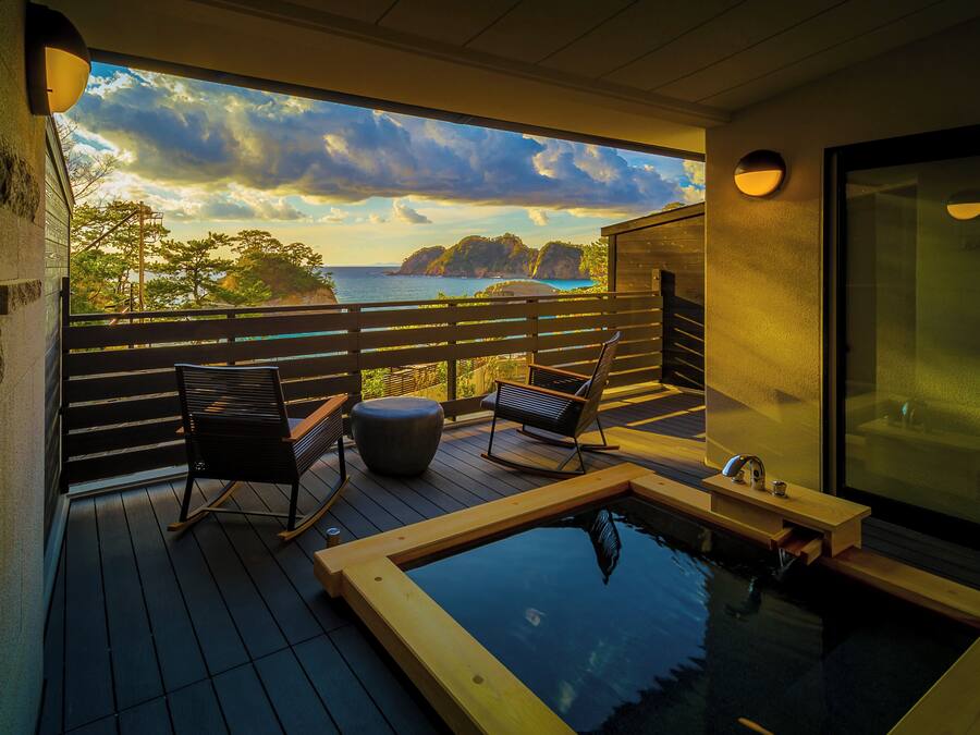 A balcony with wooden flooring, two rocking chairs, a small wooden table, and a hot tub.