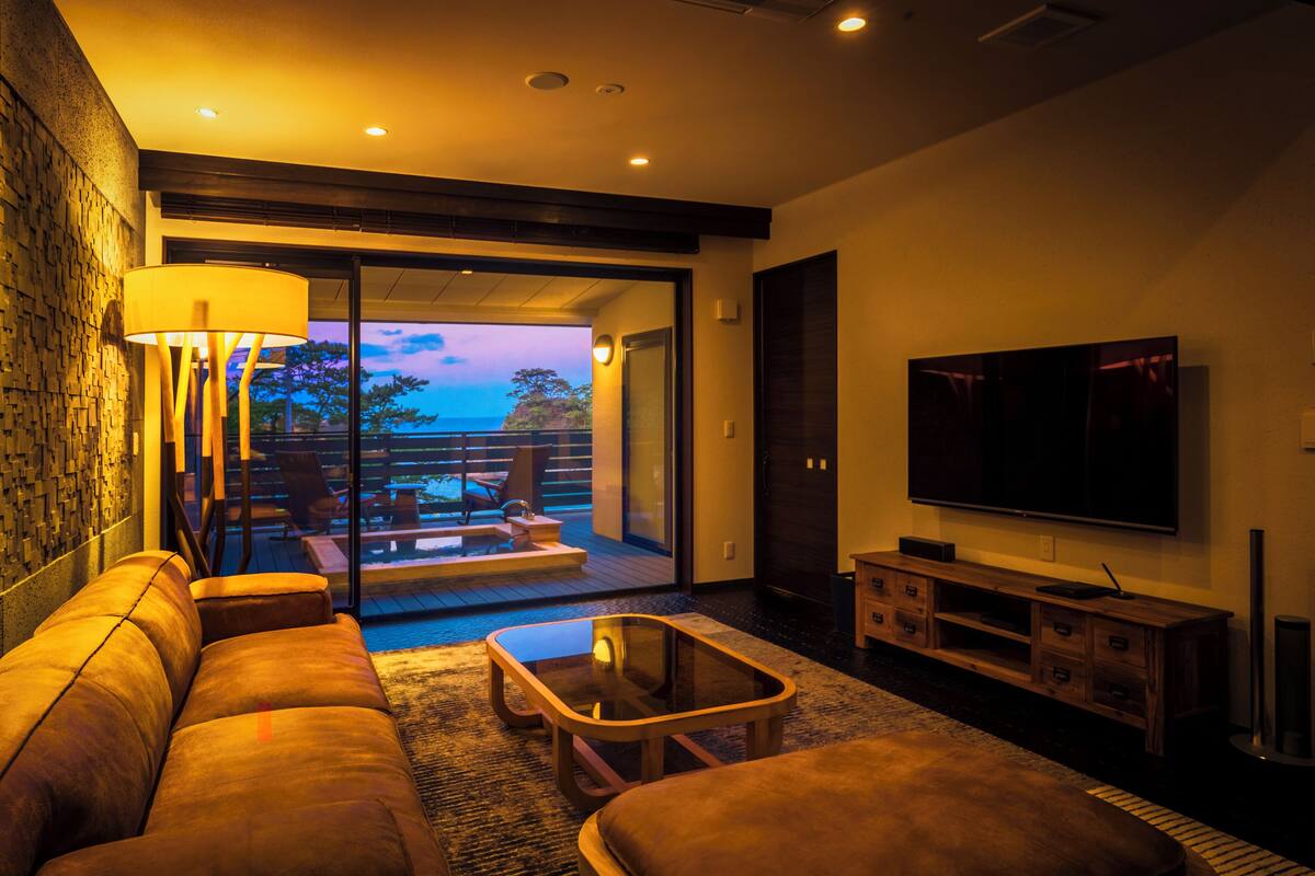 A modern living room with a sofa, coffee table, and TV. There is a balcony with a view of the sky.