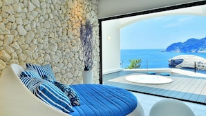 A modern bedroom with a stone wall, a large bed with blue bedding, and a view of the sea. - Mayu Futahari (Nishiizu)
