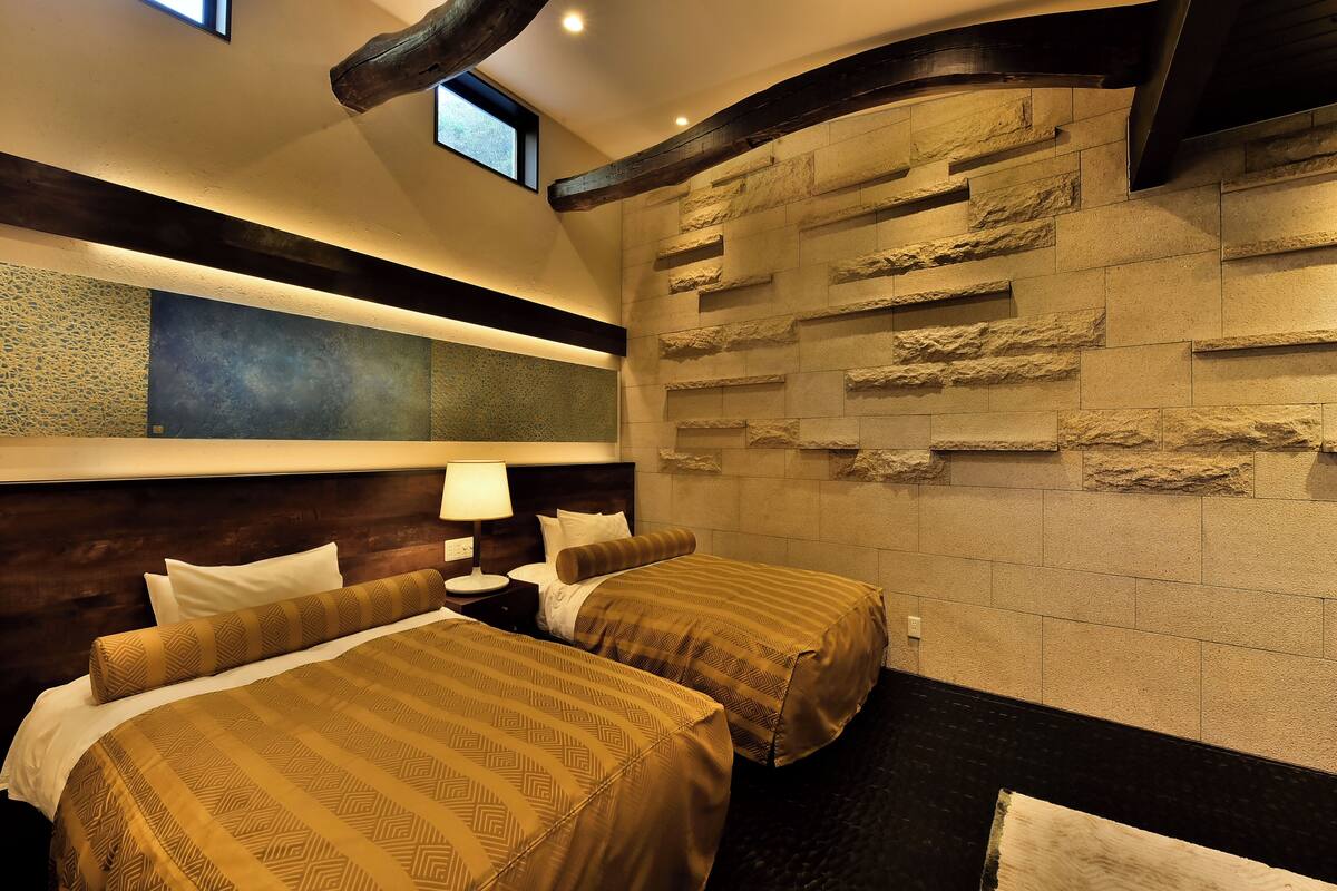 A hotel room with two beds, a nightstand, a lamp, a large mirror, and a stone wall.