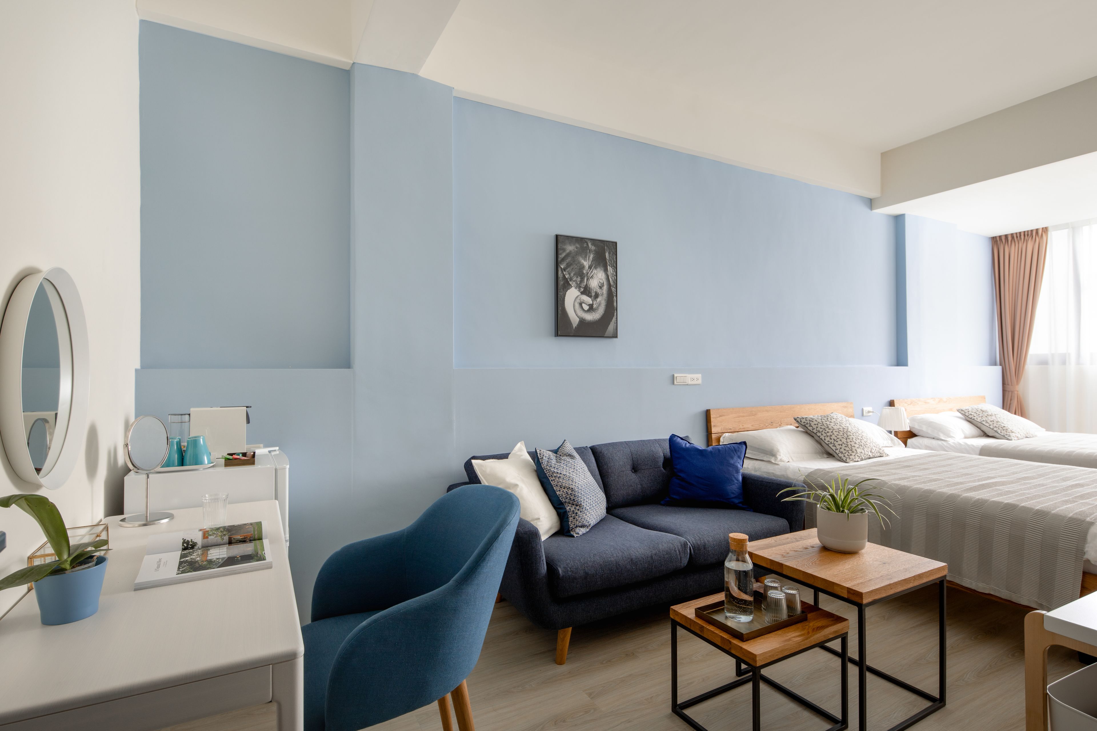 A modern hotel room with a blue accent wall, a sofa, a bed, a wooden coffee table, and a desk with a mirror and a plant.