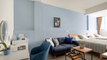 A modern hotel room with a blue accent wall, a sofa, a bed, a wooden coffee table, and a desk with a mirror and a plant.