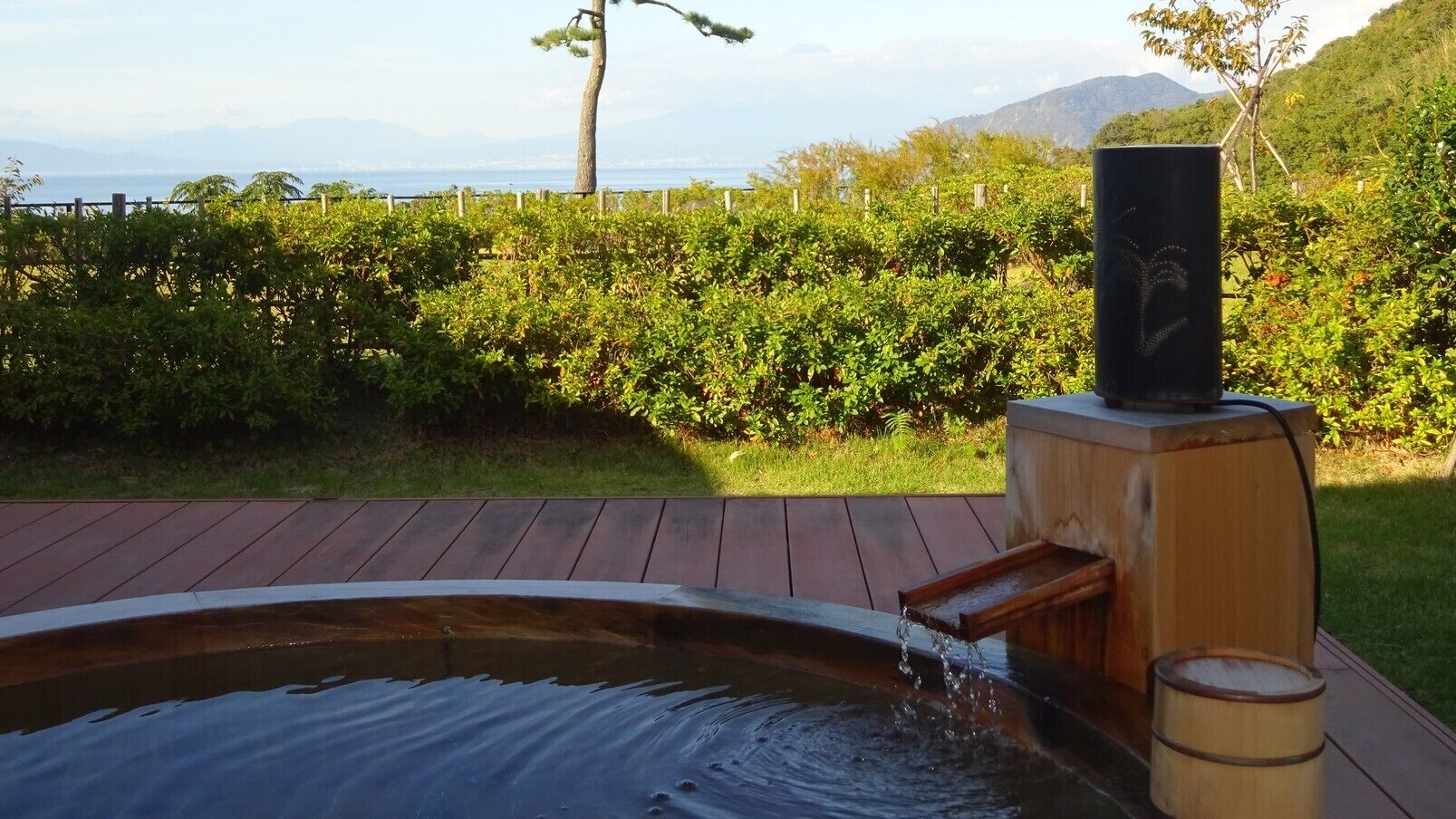 A hot tub with a waterfall feature, surrounded by a wooden deck and a hedge.
