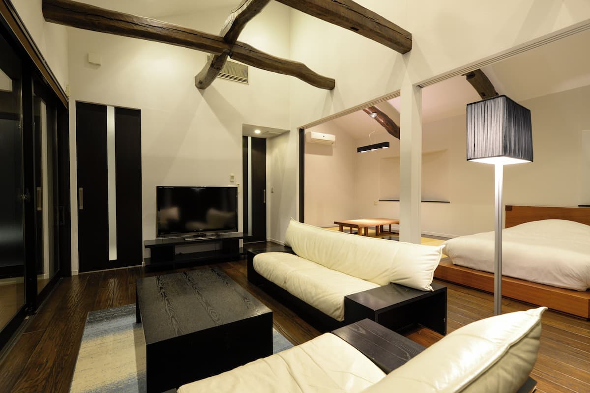 Una sala de estar moderna con un sofá, una mesa de centro y un televisor. Se aprecia una viga de madera en el techo, una lámpara y una cama al fondo.