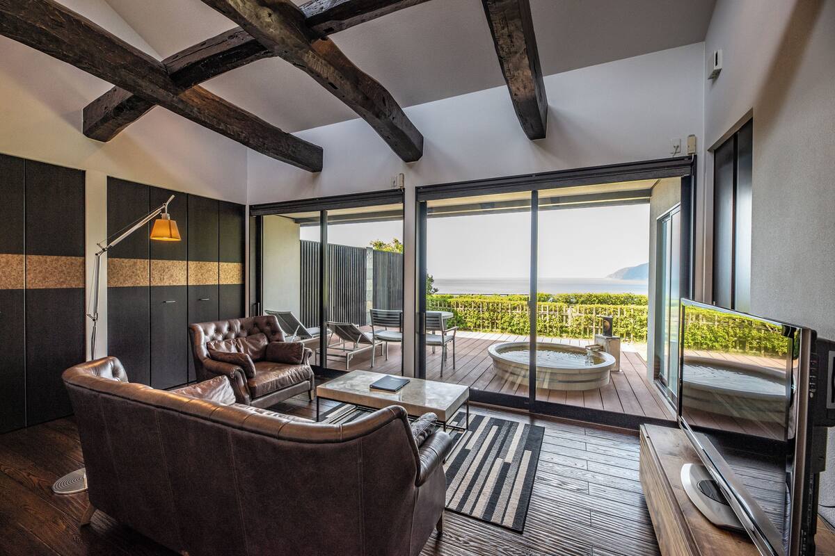 Una sala de estar moderna con sofás de cuero, una mesa de centro y un televisor. Cuenta con ventanas grandes con vistas a un viñedo y un jacuzzi en el exterior.