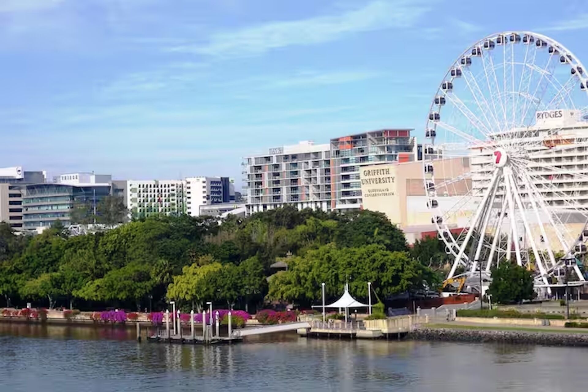 Blick auf das Wasser mit Riesenrad, einem Universitätsgebäude und einem Park mit Laube.