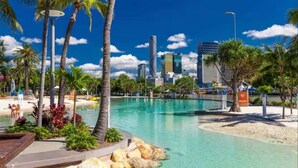 A resort pool area with palm trees, a clear blue sky, and a city skyline in the background. - Park Avenue Luxe Chic Townhouse Sleeps 9, Carpark (Auchenflower)