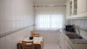 Cocina pequeña con paredes de azulejos blancos, una mesa de comedor con sillas, una ventana con cortinas translúcidas y un lavamanos empotrado.