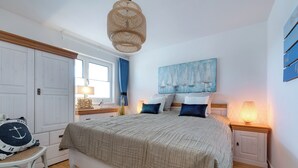 Een slaapkamer met een bed, nachtkastje, stoel, raam met gordijnen en een schilderij aan de muur.