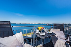 A balcony with a table set for two, overlooking a body of water with a pier in the distance. - Seestern - Elevator, Balcony, Underfloor Heating, TV, First Row Waterfront (Olpenitz)