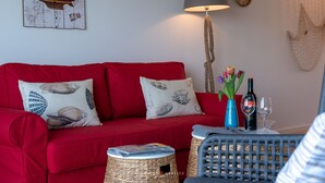 Een rode bank met decoratieve kussens, een lamp, een vaas met bloemen en een fles wijn op een bijzettafeltje.