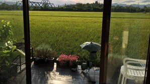 Blick auf ein grünes Feld durch ein Fenster, im Hintergrund eine Metallkonstruktion und auf der Terrasse ein Stuhl und ein Sonnenschirm.
