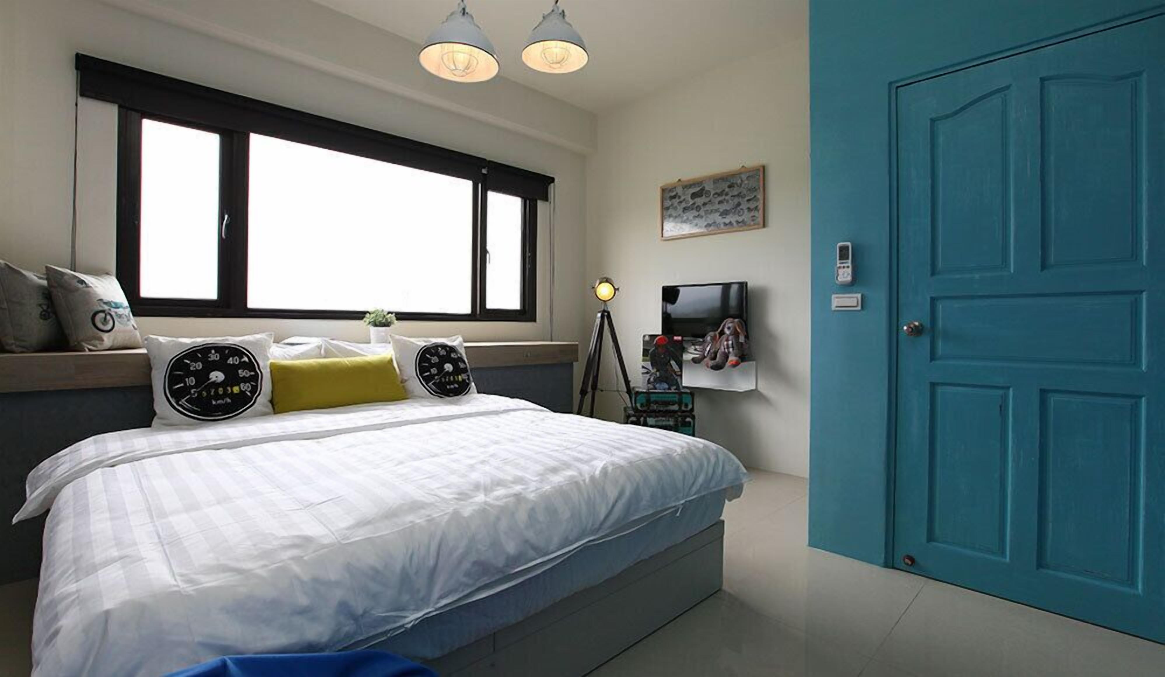 A bedroom with a large bed, a window with blinds, a desk with a lamp, a television, and a blue door.