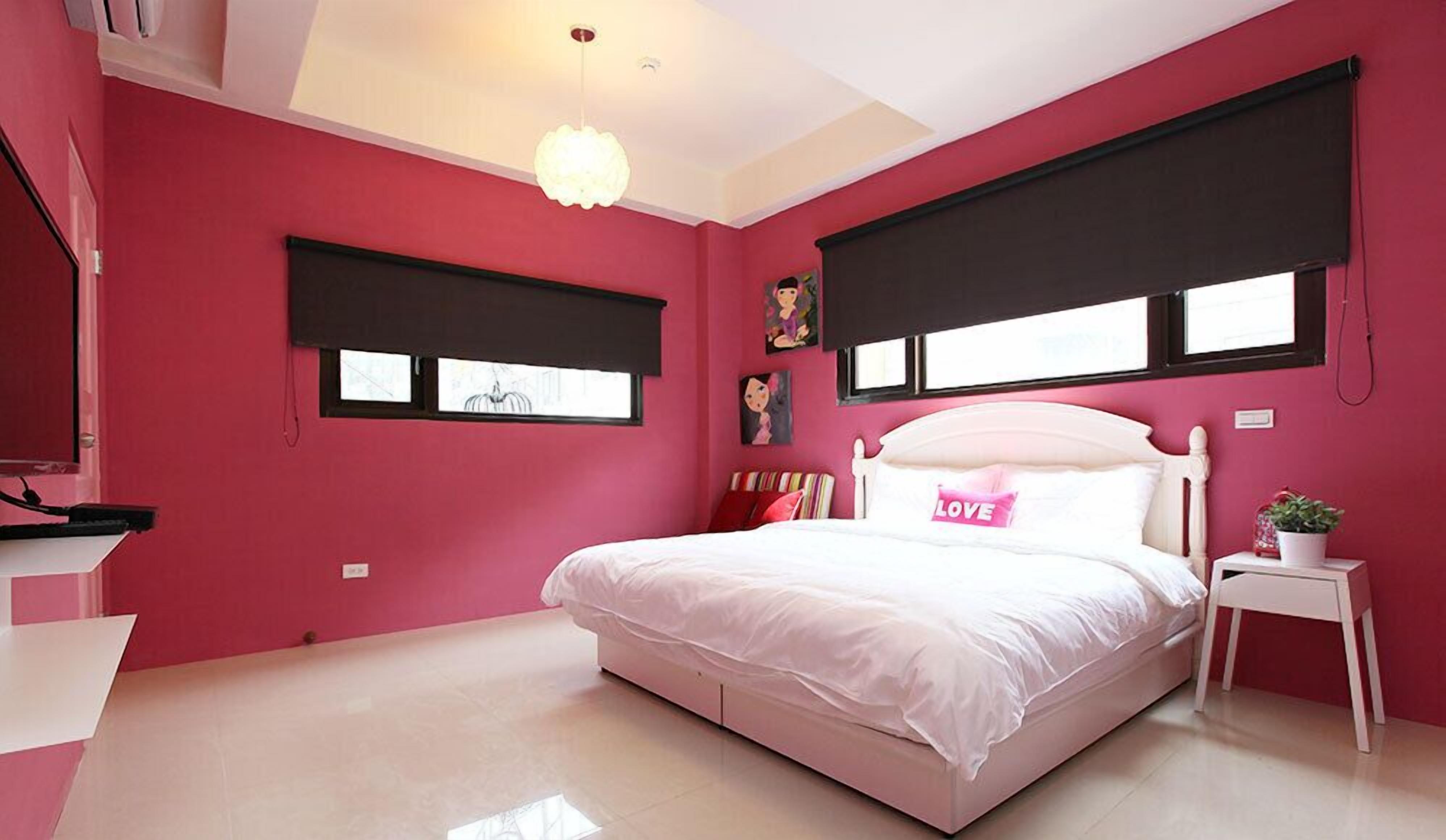 A bedroom with a pink wall, a white bed, a bedside table with a plant, and a window with a black blind.