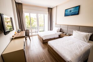 A hotel room with two beds, a TV, a desk, and a balcony with a view of palm trees.