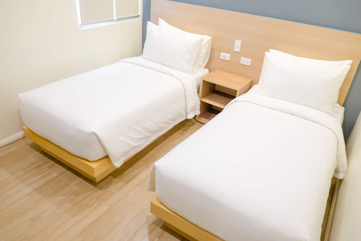 A hotel room with two single beds, wooden headboards, a small wooden nightstand, and a window with blinds.