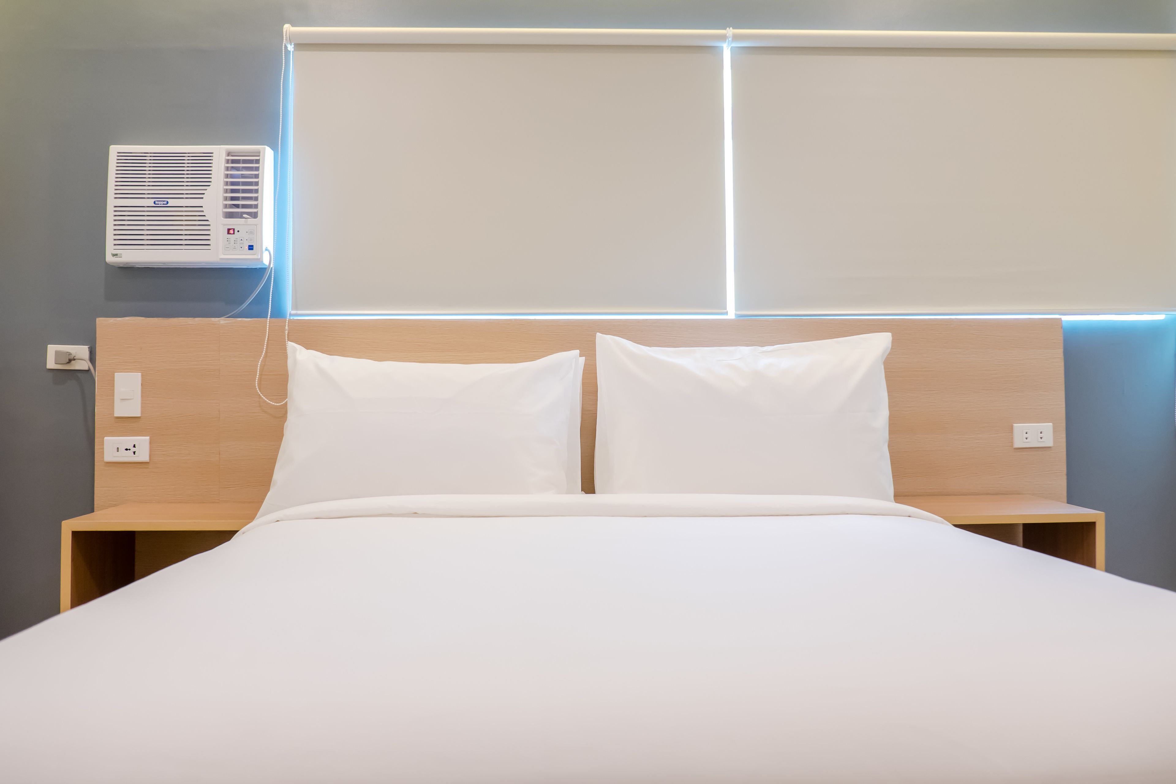 A neatly made bed with white linens, a wooden headboard, and a wall-mounted air conditioner.