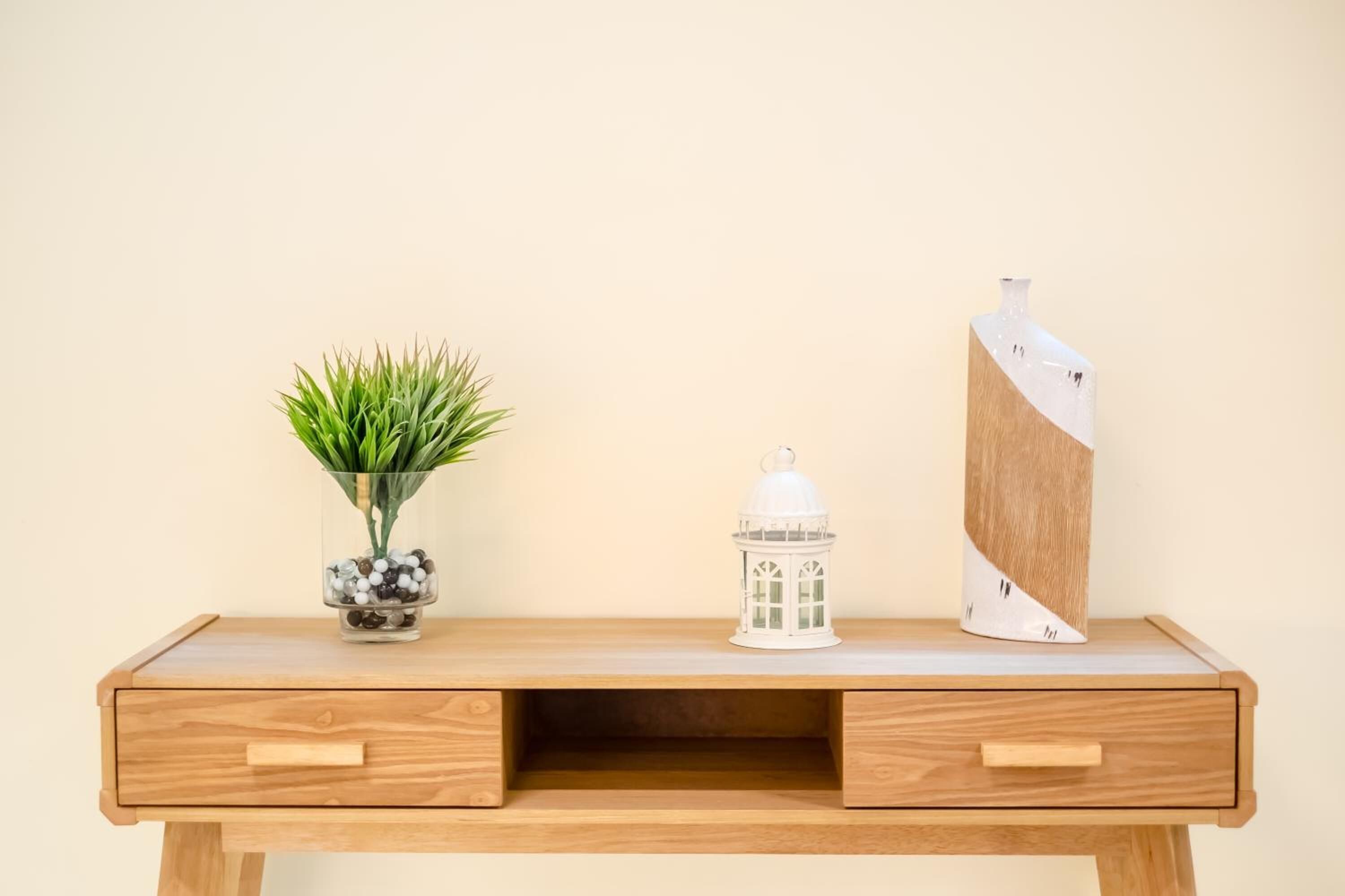 Un bureau en bois doté de deux tiroirs, un vase en verre contenant des plantes vertes, une lanterne blanche et un vase blanc orné d’un motif géométrique.