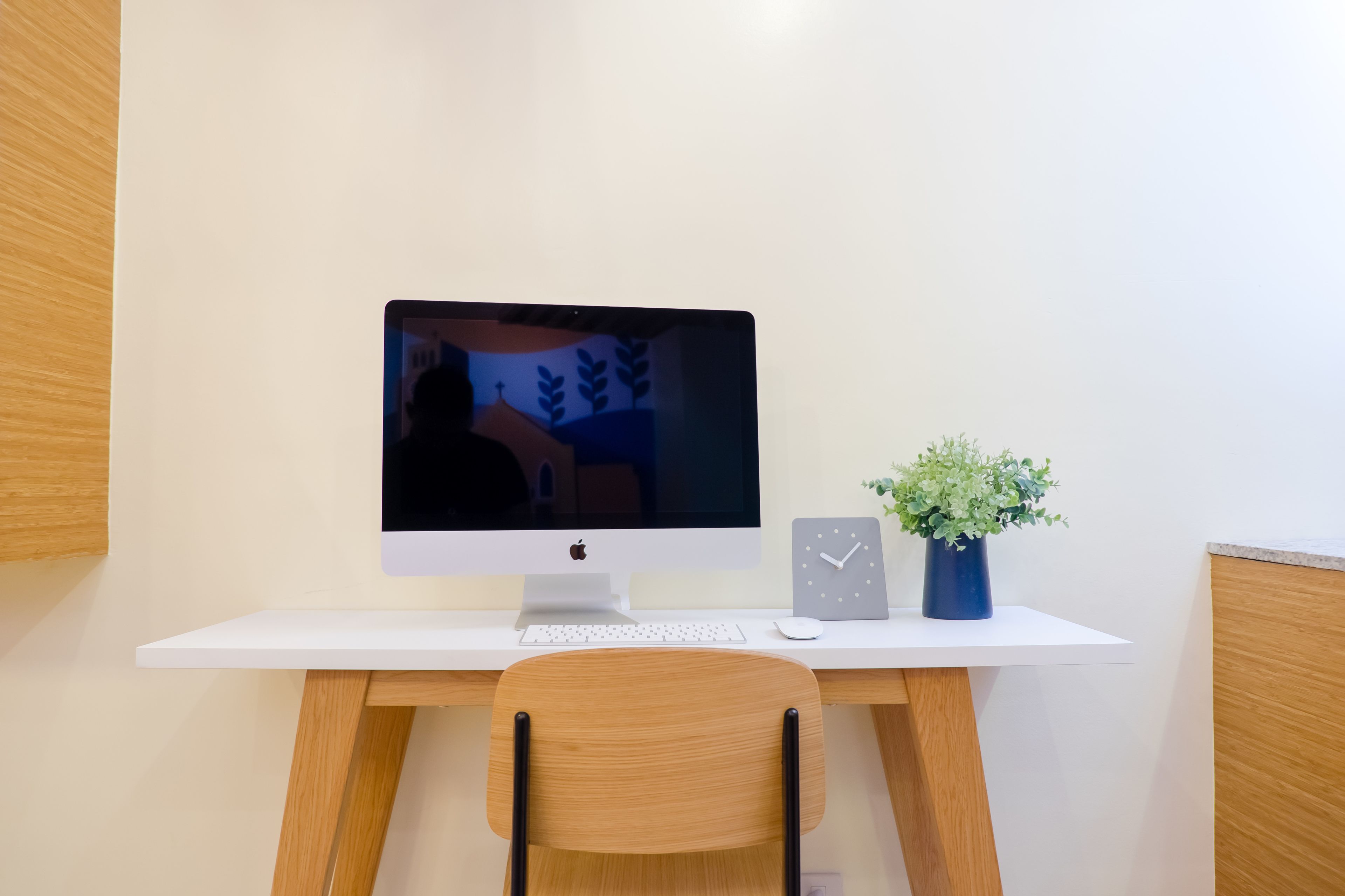 A white desk with a computer monitor, keyboard, mouse, and a clock. There is a chair in front of the desk and a small potted plant on the right side.