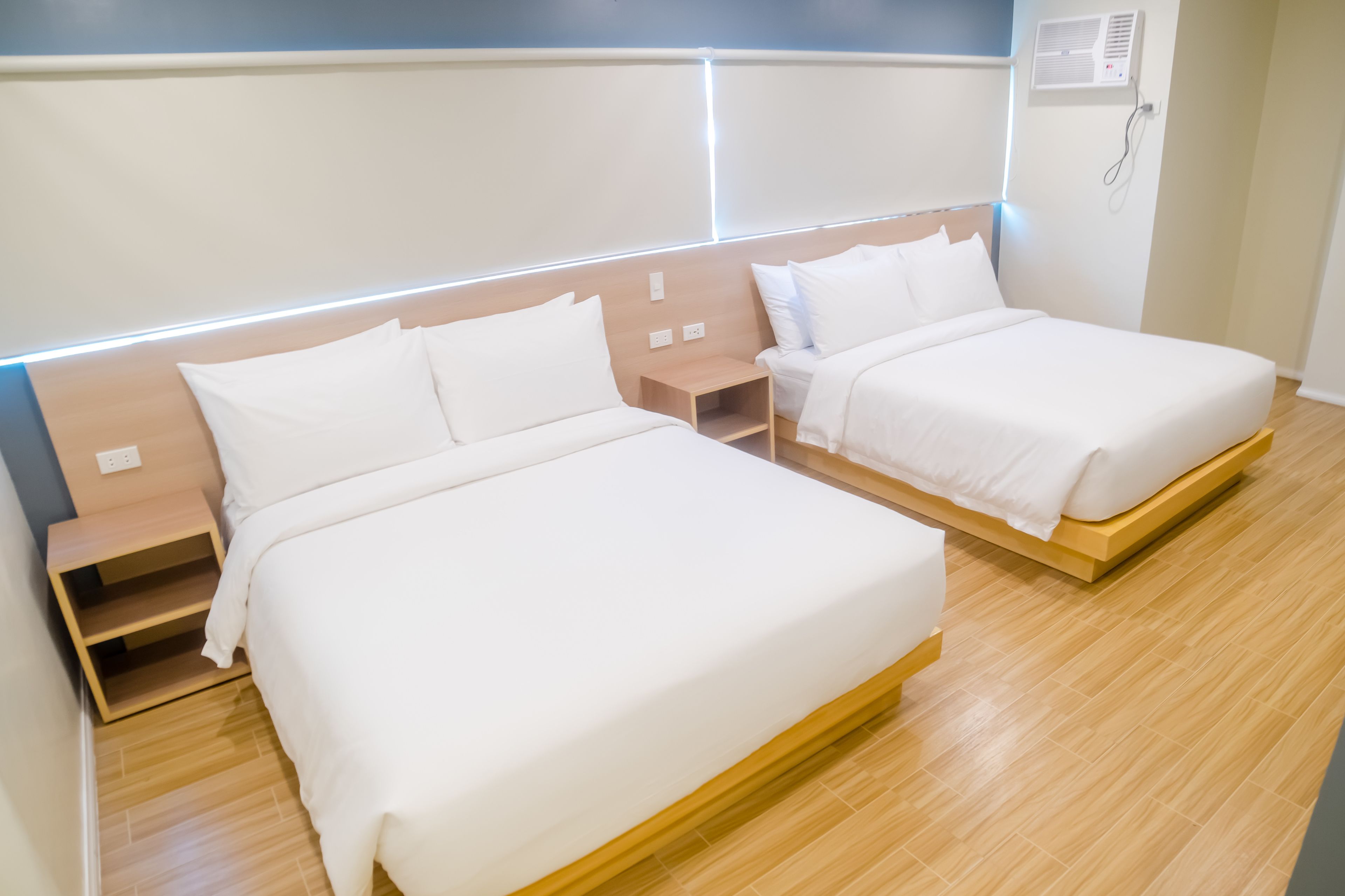 A hotel room with two beds, wooden headboards, bedside tables, and a wall-mounted air conditioner.