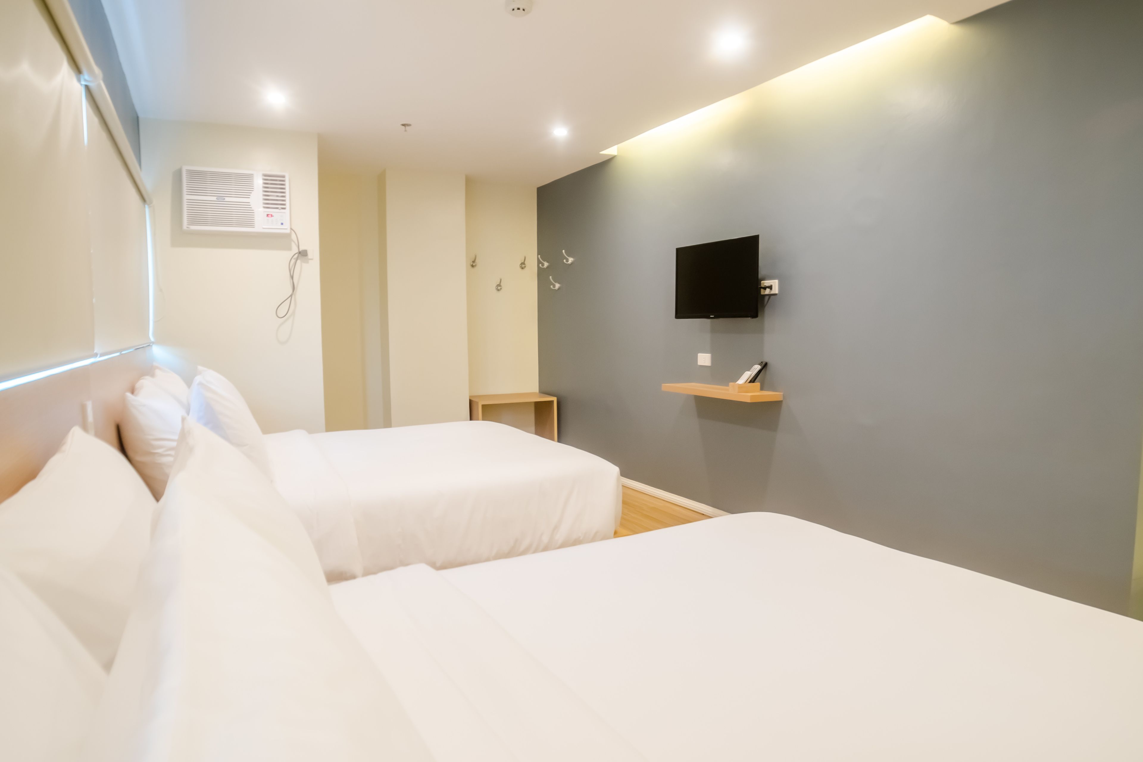 A hotel room with two beds, a TV mounted on the wall, a small wooden side table, and an air conditioning unit on the wall.