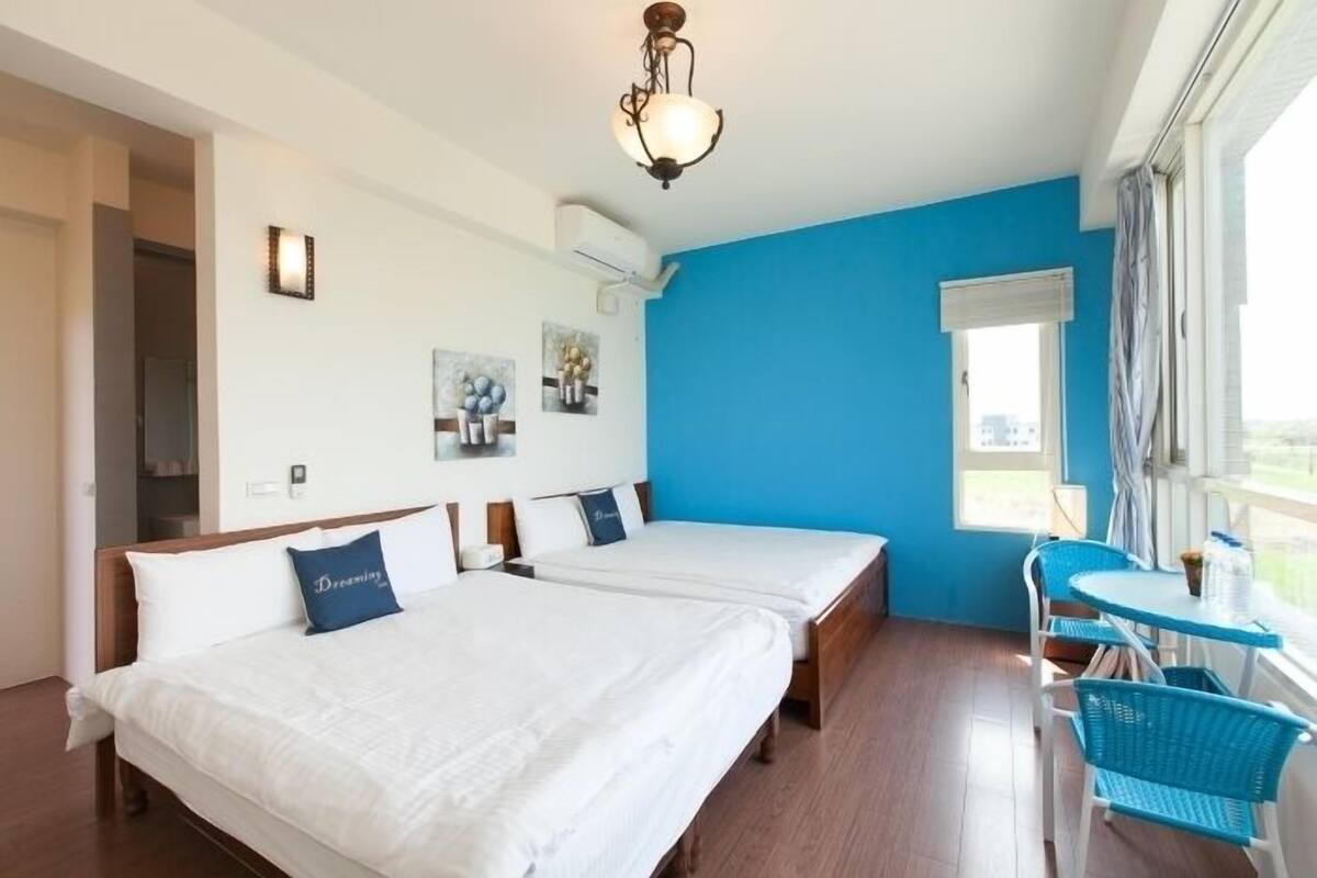 Habitación de hotel con dos camas, un área de comedor con mesa y sillas, y un gran ventanal con cortinas.