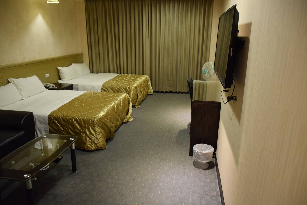 A hotel room with two beds, a sofa, a glass-top table, a trash bin, and a television.