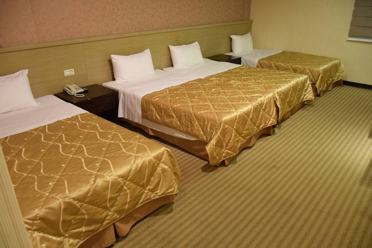 A hotel room with three single beds, each with a gold bedspread and white pillows. There is a telephone on the nightstand next to the first bed.