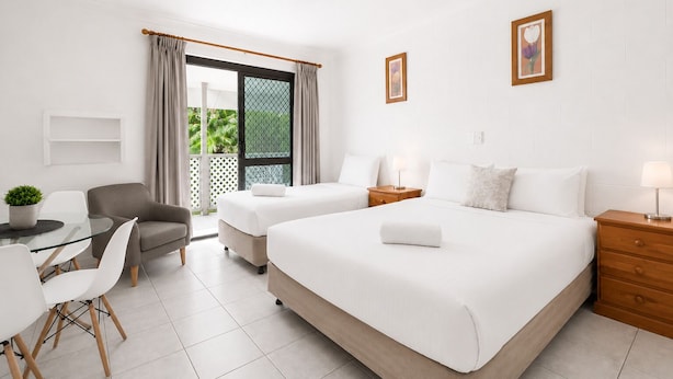 A hotel room with two beds, a desk with chairs, a ceiling fan, and a view of greenery outside.