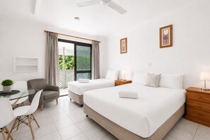 Ein Hotelzimmer mit zwei Betten, einem Schreibtisch mit Stühlen, einem Deckenventilator und Blick auf Grünfläche.