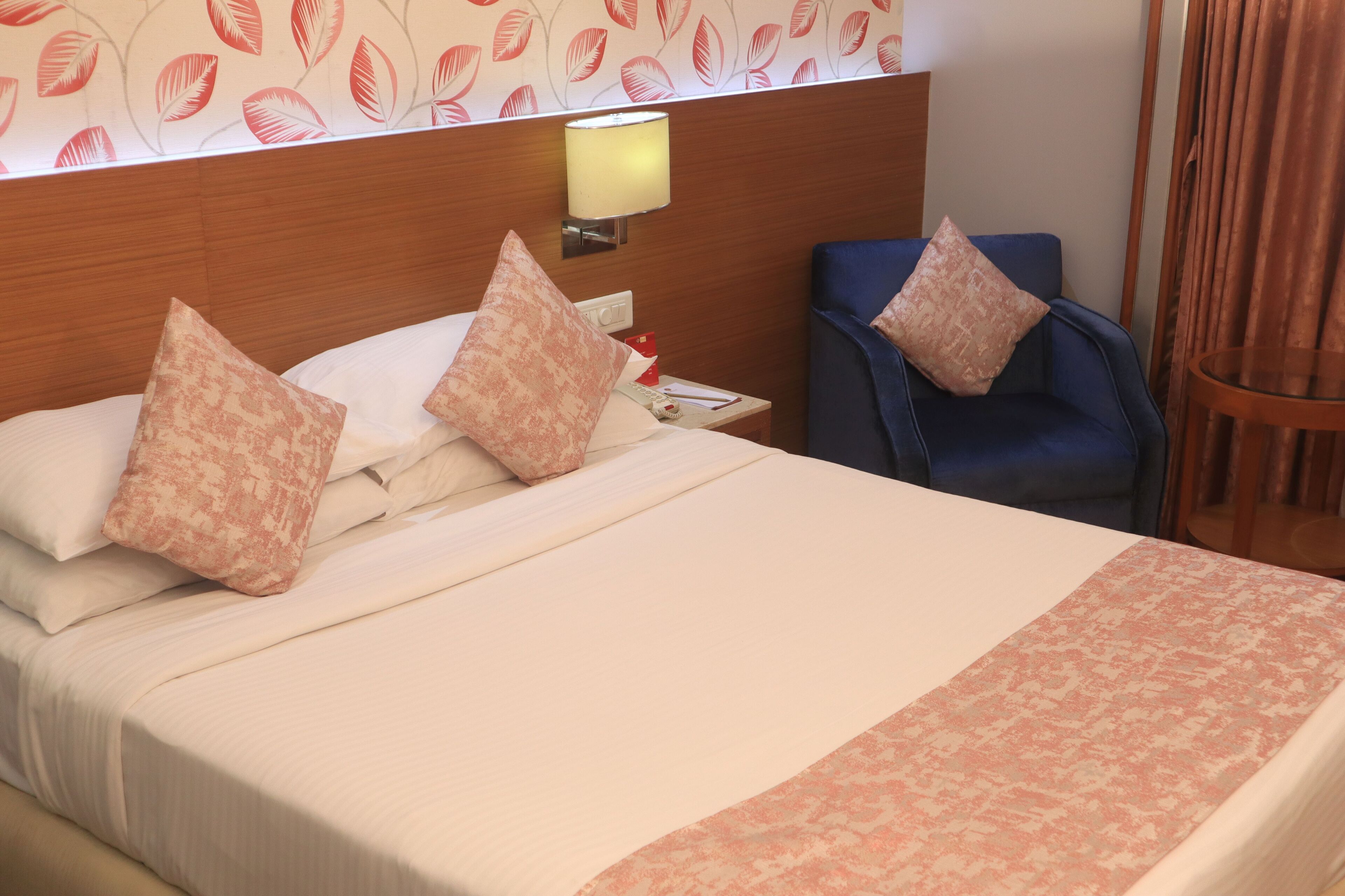 A hotel room with a bed, pillows, a nightstand, a lamp, a chair, and a curtain.