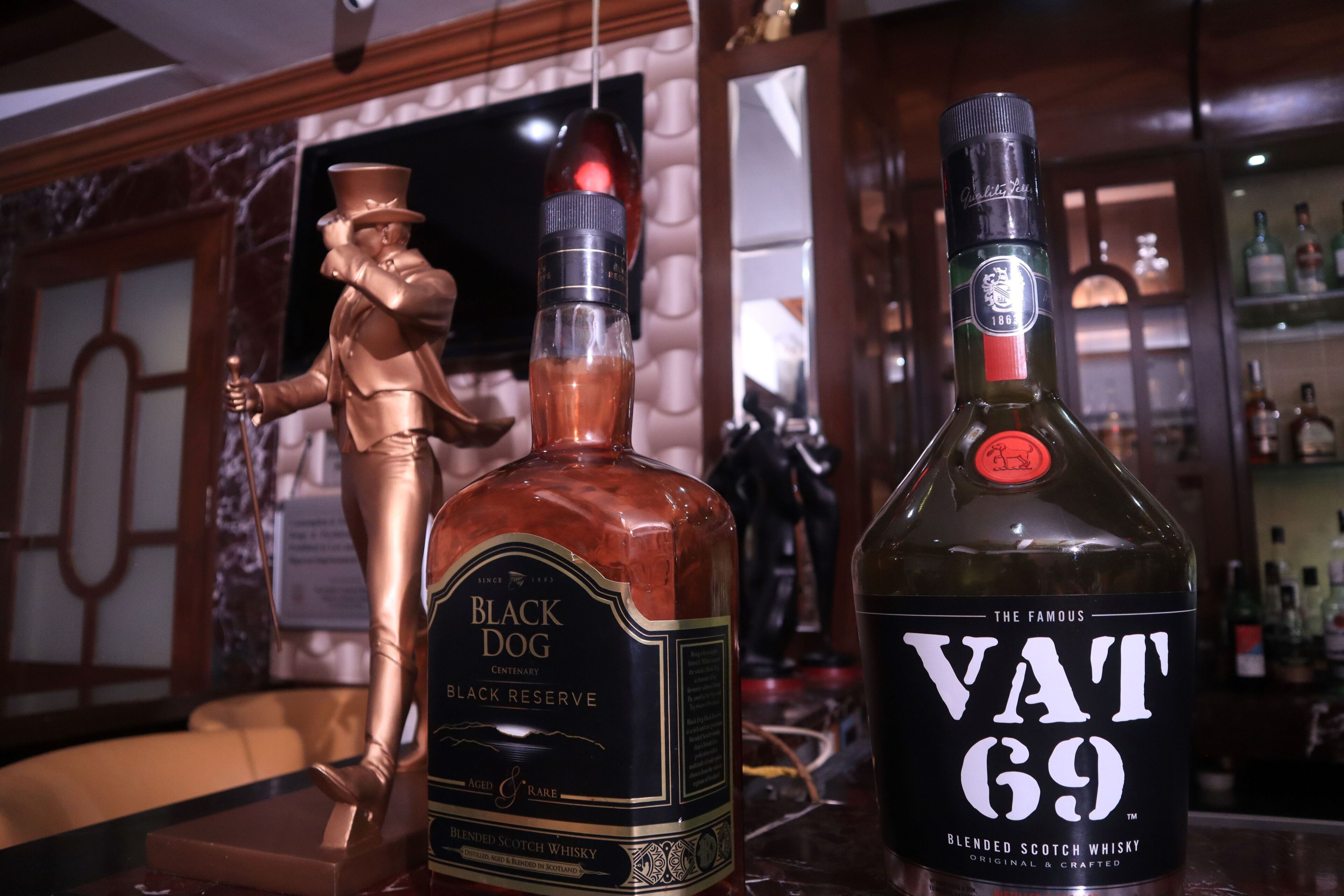 Deux bouteilles de whisky, une Black Dog Black Reserve et une Vat 69, sont posées sur le comptoir d’un bar, avec une statue en arrière-plan.