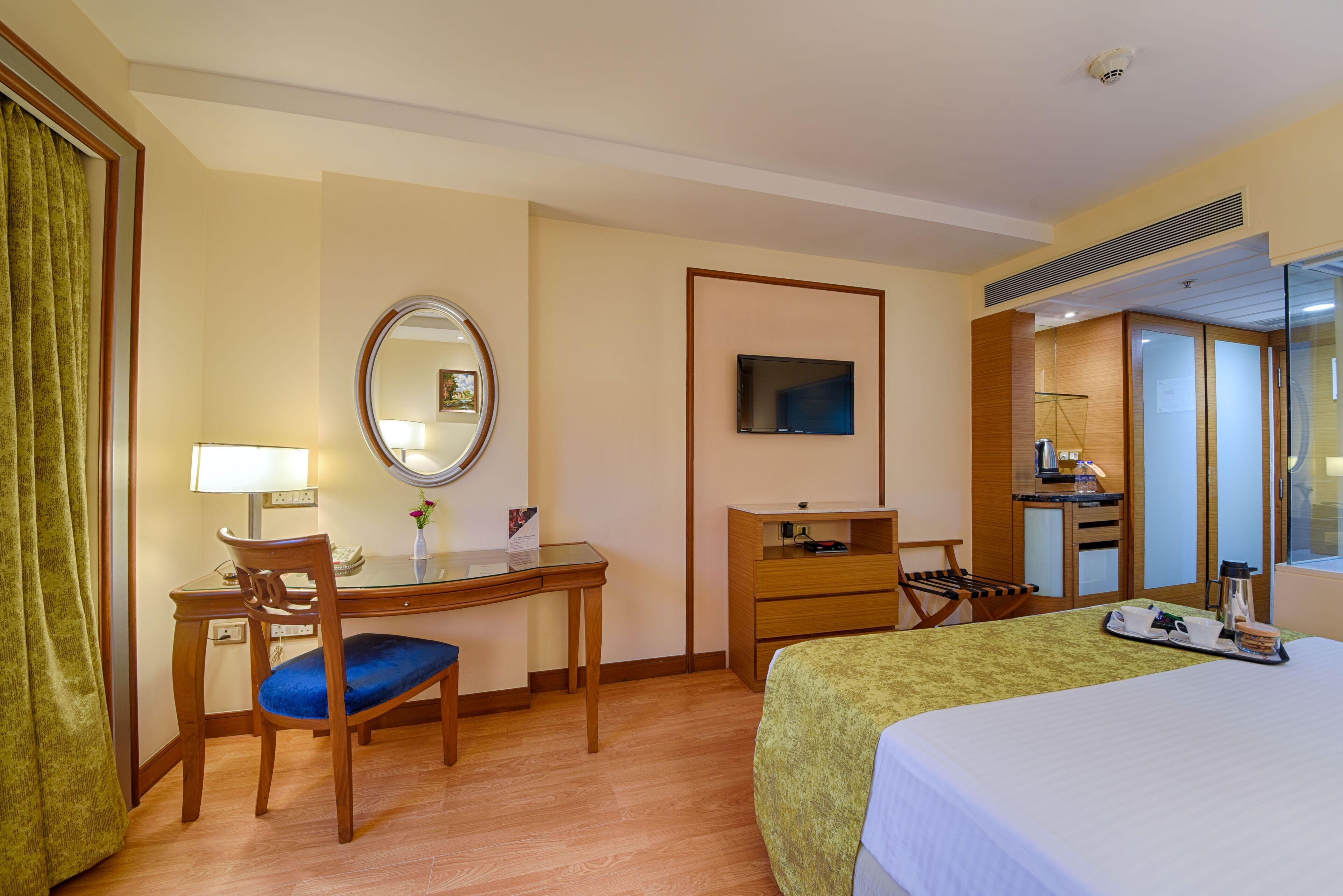 A hotel room with a bed, a desk with a chair, a mirror, a television, a small kitchenette, and a bathroom visible through a glass door.