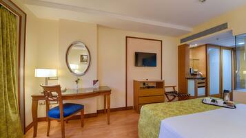 A hotel room with a bed, a desk with a chair, a mirror, a television, a small kitchenette, and a bathroom visible through a glass door.