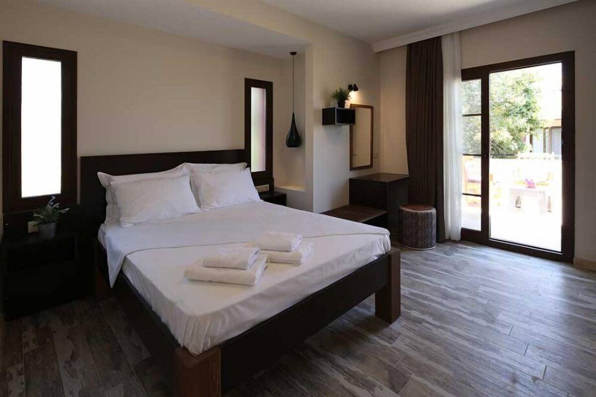 A bedroom with a large bed, a bedside table, a nightstand, a mirror, a potted plant, and a view of the outdoors through a sliding door.