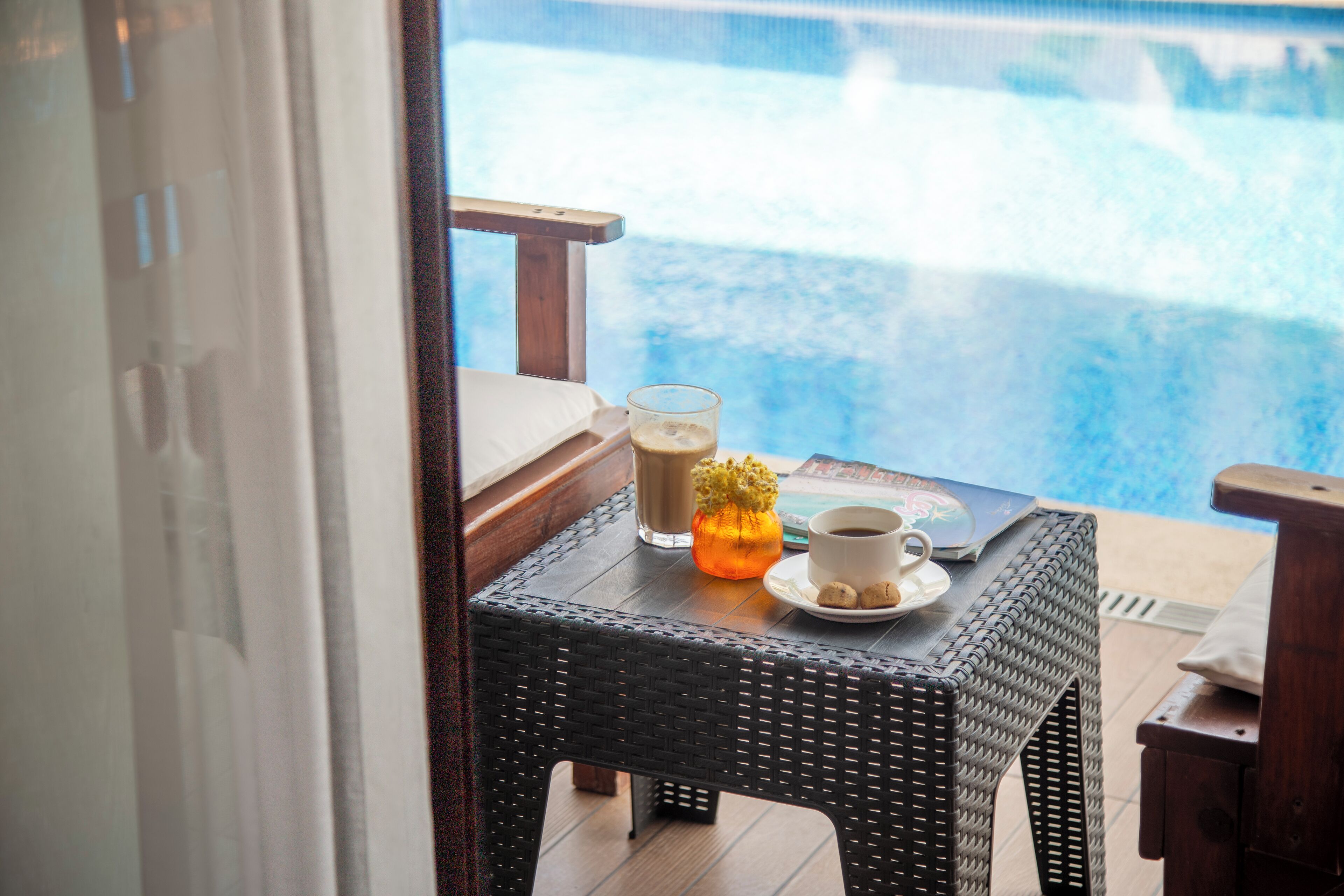 Una mesa de mimbre con un vaso de café helado, una taza de café y un pequeño florero naranja junto a una piscina.