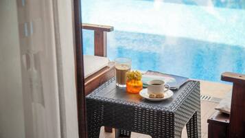Una mesa de mimbre con un vaso de café helado, una taza de café y un pequeño florero naranja junto a una piscina.