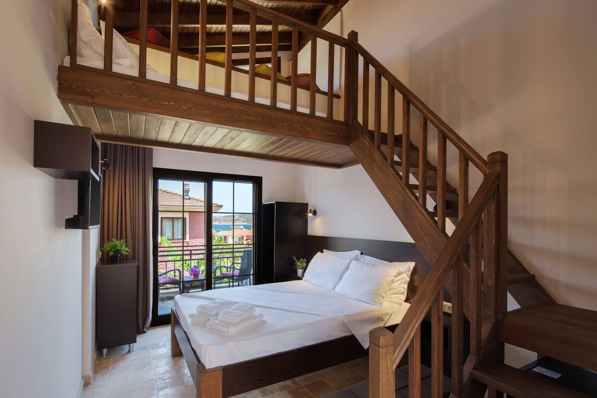 A bedroom with a large bed, wooden staircase, and a balcony with a view of buildings.