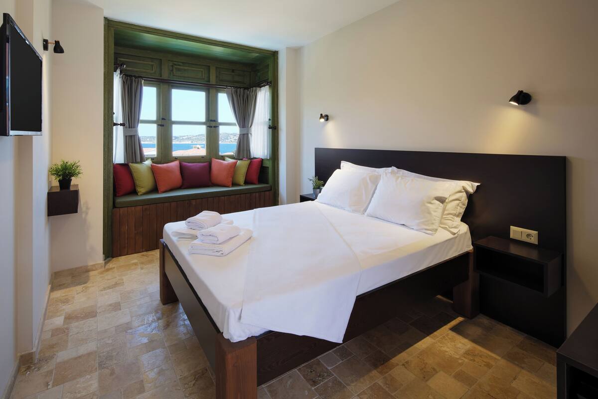 A neatly made bed with white linens, a window seat with colorful cushions, and a view of the sea.