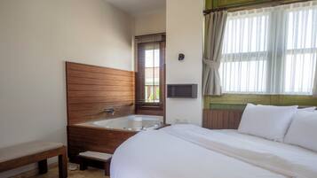 A bedroom with a large bed, a wooden headboard, a bench, a bathtub, and a window with curtains.