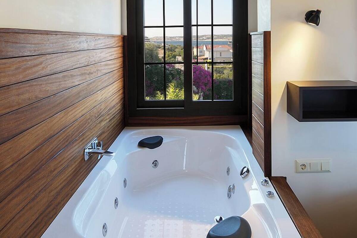 A modern bathroom with a large bathtub, wooden paneling, a window with a view, and a wall-mounted light fixture.