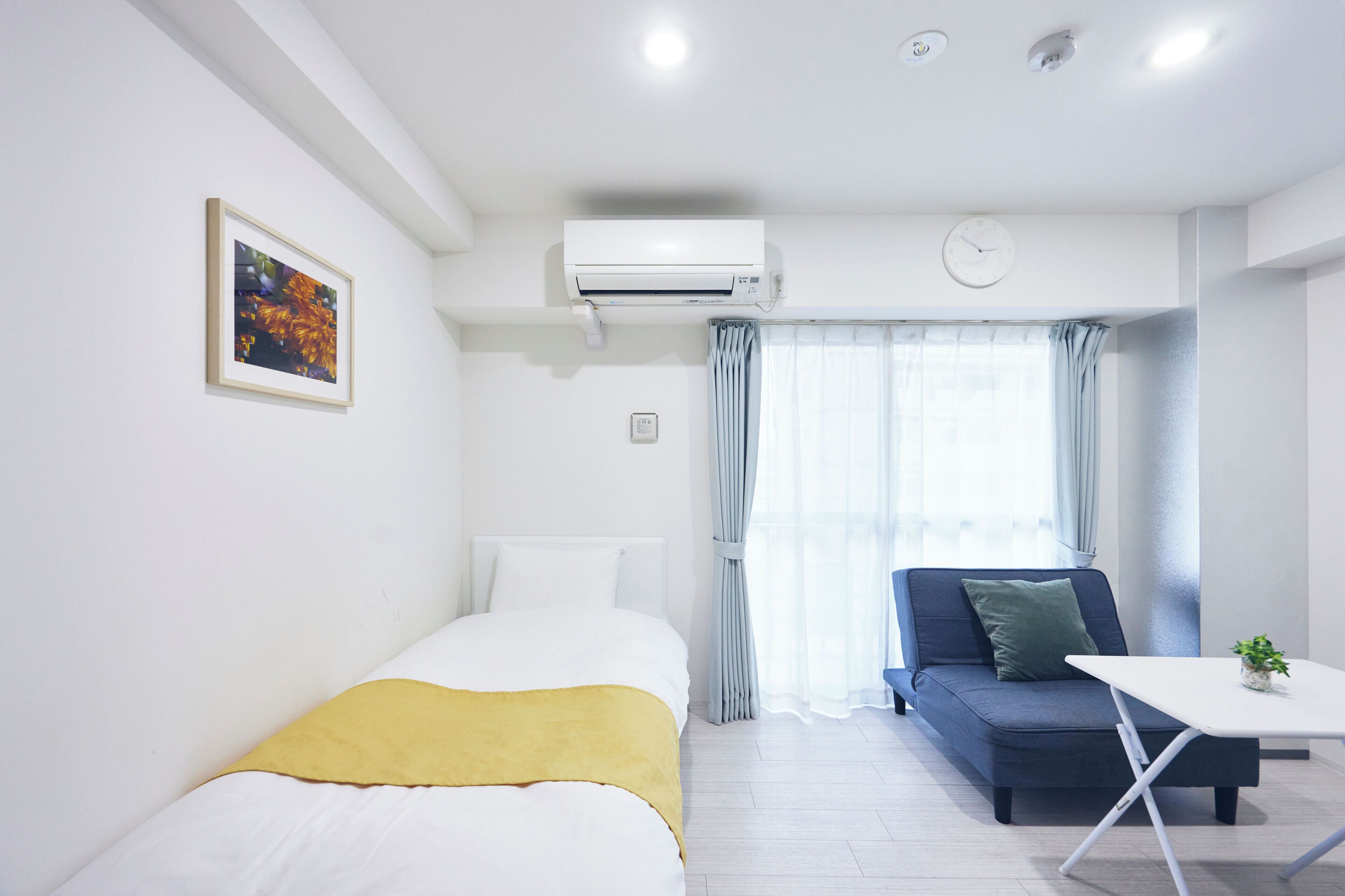 A modern hotel room with a bed, a sofa, a small table, an air conditioning unit, a clock, and a window with curtains.