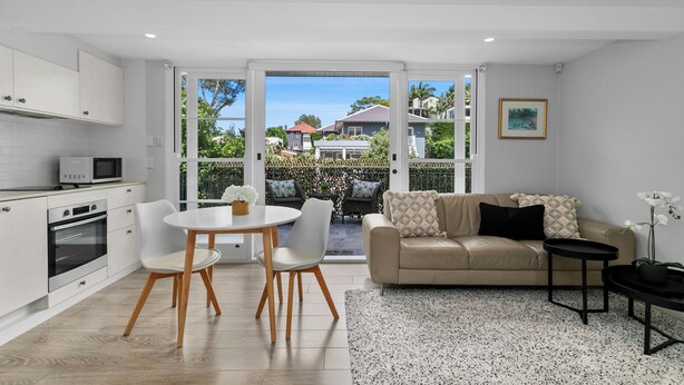 A modern kitchen with white cabinets, a dining area with a round table and chairs, a beige sofa, and a view of the neighborhood through large glass doors. - Balmoral Cabana - only 150m to Balmoral Beach (Mosman)