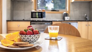 Une table de petit-déjeuner avec un bol de fraises et des tranches d’orange sur une assiette, un verre de jus d’orange et une table en bois dans une cuisine équipée de placards et d’un four à micro-ondes.