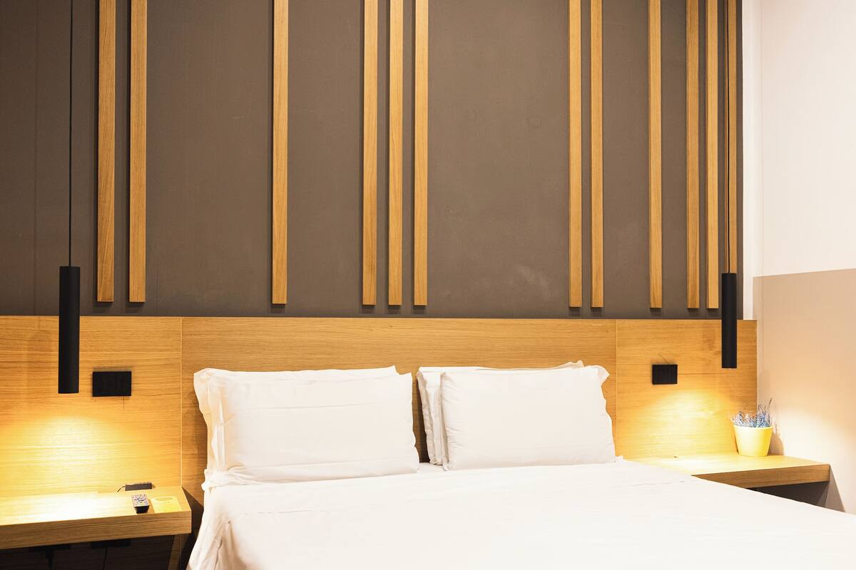 A hotel room with a wooden headboard, two white pillows, a bedside table with a lamp, and a small potted plant.