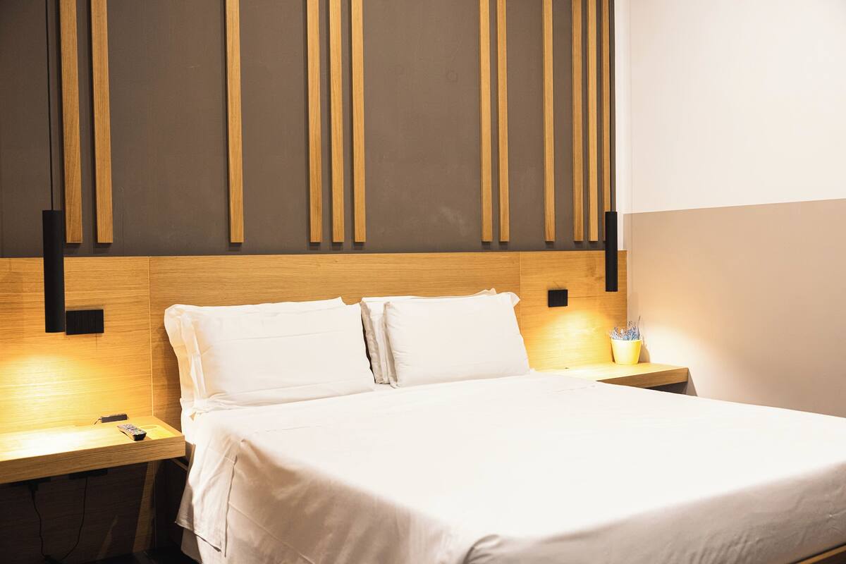 A neatly made bed with white linens in a modern room with wooden paneling and a nightstand with a lamp and a remote control.