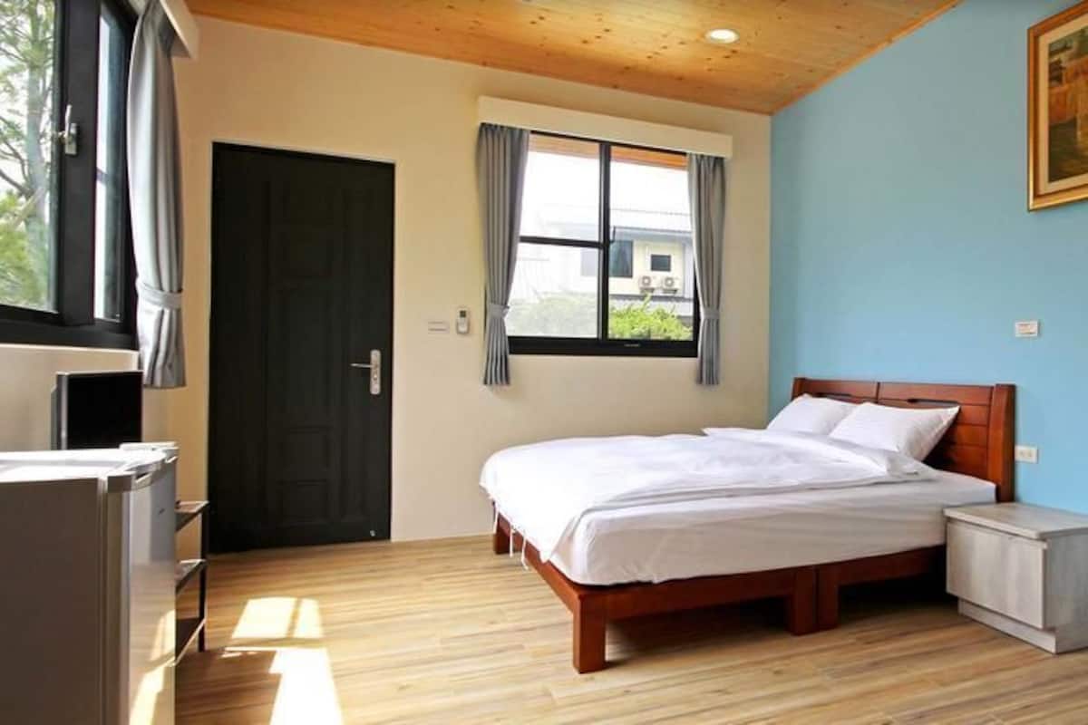 A bedroom with a wooden bed, a bedside table, a window with curtains, and a painting on the wall.